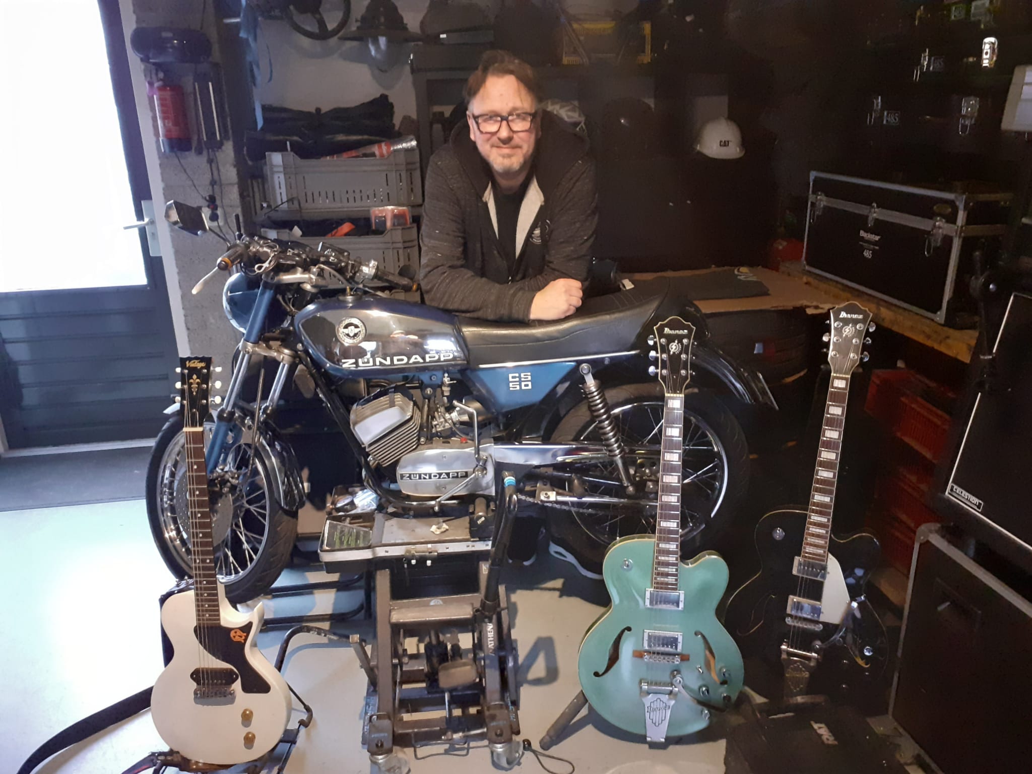 Scheuren met Perry van de Ven op het WMW
Al bij binnenkomst zie je de gitaren en Zündapp brommer staan. Dat is rocker Perry van de Ven ten voeten uit. Hij begon als drummer in ‘de keet’ bij Wientjes. Samen met de vriendengroep speelden ze covers en van alles en nog wat. “Van een optreden is het nooit gekomen,” zegt Perry. “We waren te onzeker.” Vandaar dat u nooit van de band ‘The Silence’ heeft gehoord. Daarna ging het snel met de muziekcarrière van Perry. Het is bijna te veel om allemaal op te noemen, maar hier een overzicht; elektrische gitaar bij een rock- en popkoor in Venhorst, bij ‘Take Death’ zanger en gitarist en als hoogtepunt een optreden bij Giel Beelen op 3FM met de band ‘Dawn’, later ‘American Idiots’ een coverband. Kortom; we hebben hier te maken met een doorgewinterde rocker!
Fantic walm
Perry is ook een brommerfanaat. Zijn eerste brommer was een Fantic uit Italië. Deze was ook echt Italiaans: meer repareren dan rijden. Nu gaat het beter: 50% rijden en 50% sleutelen op zijn Zündapp. Samen met zijn medebrommerkameraden vormen ze ‘Wanne Walm’, een brommertoerclub. “Prachtig vind ik dit, slappe kwats en veul lachen,” zegt Perry, “vooral als we met zijn allen een doodlopend weggetje in rijden, dan is het walmend chaotisch.” Af en toe wil Perry nog wel eens echt scheuren op zijn, hoe is ’t mogelijk, Zündapp. Onlangs in Mill lag hij plat in een bocht en reed bijna de wieldoppen af van een politieauto. De politieagente keek even meewarig en dacht: weer zo’n oude gek.
485 zonder bril
In zijn nieuwe band 485 (Four Eight Five) speelt Perry covers van rock en punkrock met een eigen draai. Samen met Linda Jacobs (ook WMW-artiest) en drie andere bandleden hebben ze een debuut gehad met hun optreden op de Wanroijse kermis.
Lees het hele interview op www.facebook.com/muziekverenigingstjanwanroij