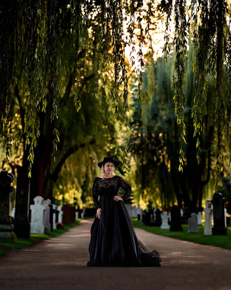 “Framed by Memory”
Under the curtain of willow branches, she stood where the path narrowed and the light softened, as if the world itself had decided to whisper.
Gravestones lined the road like quiet listeners, their names worn smooth by time and weather. They did not frighten her. Tonight, they felt like witnesses—keepers of stories that refused to vanish. The air smelled of earth and leaves, and dusk slipped between green and gold, settling gently on her shoulders.
Her black dress pooled at her feet like spilled ink, elegant and heavy with meaning. It was not mourning she wore, but remembrance. Each step she had taken to this place carried the echo of laughter, loss, love, and becoming. The hat shaded her eyes, but not her resolve.
She placed a hand on her hip, steady and sure, and breathed in. The willows swayed above, their long fingers brushing the air as if blessing her courage. Somewhere behind her, the past waited. Somewhere ahead, the future opened.
She was not trapped between the stones—she was framed by them.
And as the last light of day slipped through the trees, she understood: stories do not end where names are carved. They continue in the living, in the brave choice to stand tall, and in the quiet power of remembering who you are—even when surrounded by silence.