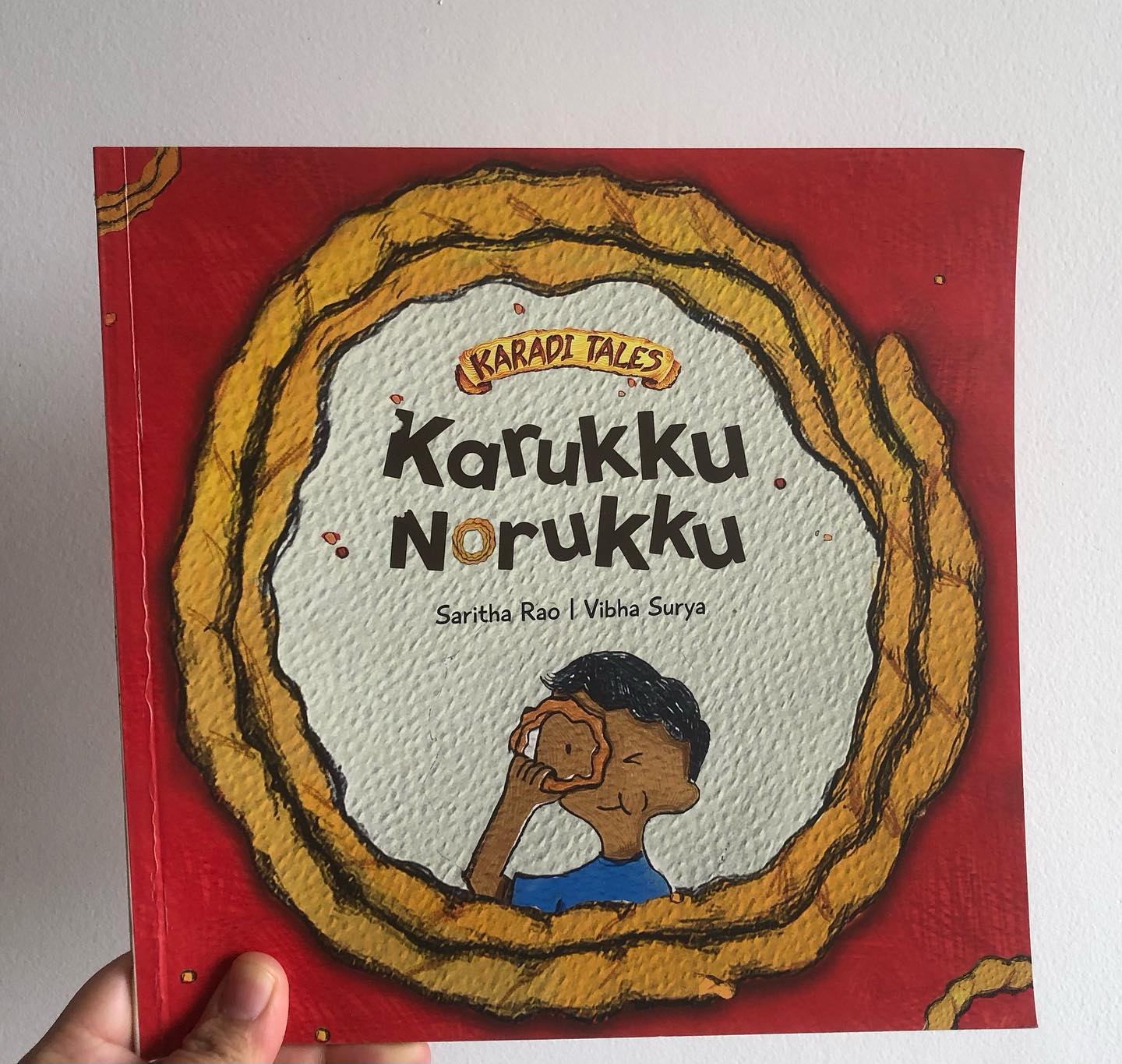 By Saritha Rao
Illustrated by Vibha Surya
Published by Karadi Tales
At first glance, Karukku Norukku is a delightful story filled with lighthearted moments. The protagonist, Guru, loves murukkus and has but one wish: to enjoy them in peace without the obligation to share. He devises a clever plan to hide a packet of murukkus in his bag, avoiding the need to share them with others. However, as the story unfolds, Guru’s plan is continuously thwarted, and despite his efforts, he is unable to eat even one murukku.
The story concludes with a seemingly fortunate turn of events as Janmashtami arrives, a day when fried snacks like murukkus are plentiful, and all is well. On the surface, it feels like a happy ending.
But let’s look at some plot points if I may.
Firstly, the central theme of the book revolves around Guru’s reluctance to share. The narrative implies that because of Guru’s unwillingness to share, he has to go into a lot of trouble to eat even one murukku. This moral feels heavy-handed and counterproductive. Teaching children to share should stem from fostering an understanding of the joy it brings, not through unnecessarily punitive consequences.
Secondly, the use of language in the book is problematic. The attempt to incorporate colloquial phrases—presumably to reflect South Indian speech—comes off as an exaggerated stereotype. The notion that “We all South Indians/Indians in general speak like this only no?!” is both inaccurate and reductive. Mainstreaming these phrases diminishes the potential for authentic representation and perpetuates unwarranted caricatures of regional speech.
The redeeming aspect of Karukku Norukku lies in its illustrations. Vibha Surya’s art is creative and detailed, bringing a quirky charm to the book that elevates its visual appeal. The artwork is indeed splendid and adds an engaging layer to the story, making it visually stimulating for young readers.
Appropriate for children aged 2 and above.