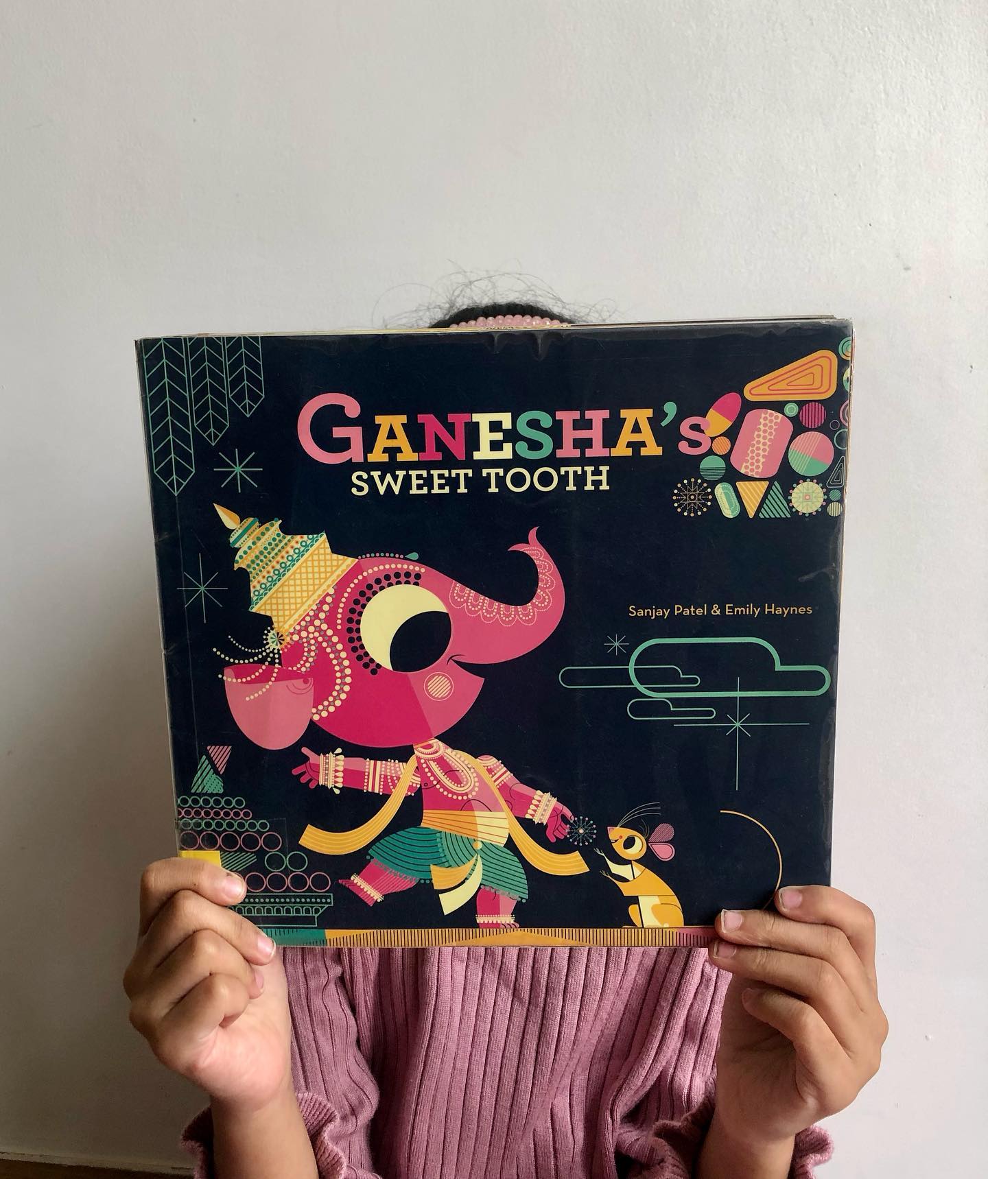 Ganesha’s Sweet Tooth
By Sanjay Patel and Emily Haynes
Indian mythology can be a tricky topic to dive into with kids—it’s often full of heavy themes, unbelievable stories, over-the-top characters, and more drama than a primetime TV show. Add in the occasional dose of epic violence, and it’s not exactly your go-to bedtime material. That’s why Ganesha’s Sweet Tooth was such a refreshing delight!
Here, Ganesha isn’t a larger-than-life god—he’s just a candy-loving kid with an insatiable sweet tooth. Together with his loyal and hilarious buddy, Mr. Mouse, Ganesha is on a sugary rampage until he encounters the Super Jumbo Jawbreaker Laddoo. Naturally, this laddoo turns out to be his match, breaking Ganesha’s tusk in the process.
In classic tantrum style, Ganesha flings the tusk in frustration—only for it to land on Vyasa, who, with some quick thinking, transforms the situation into the start of a legendary partnership. Cue the creation of the Mahabharata!
We all know the story of Ganesha scribing the Mahabharata, but this retelling brings it to life with humor and creativity. It’s age-appropriate, clever, and even sneaks in some SEL lessons. From Ganesha’s temper tantrum to his eventual “turn lemons into lemonade” moment, there’s a subtle message about adapting to life’s curveballs.
The illustrations are a visual treat—think bold, modern colors and graphics that feel straight out of a futuristic video game. This playful reimagining of Ganesha and his ever-patient “manager,” Mr. Mouse, had us hooked.
Perfect for kids aged 4+ and their grown-ups who love a fresh twist on old classics.