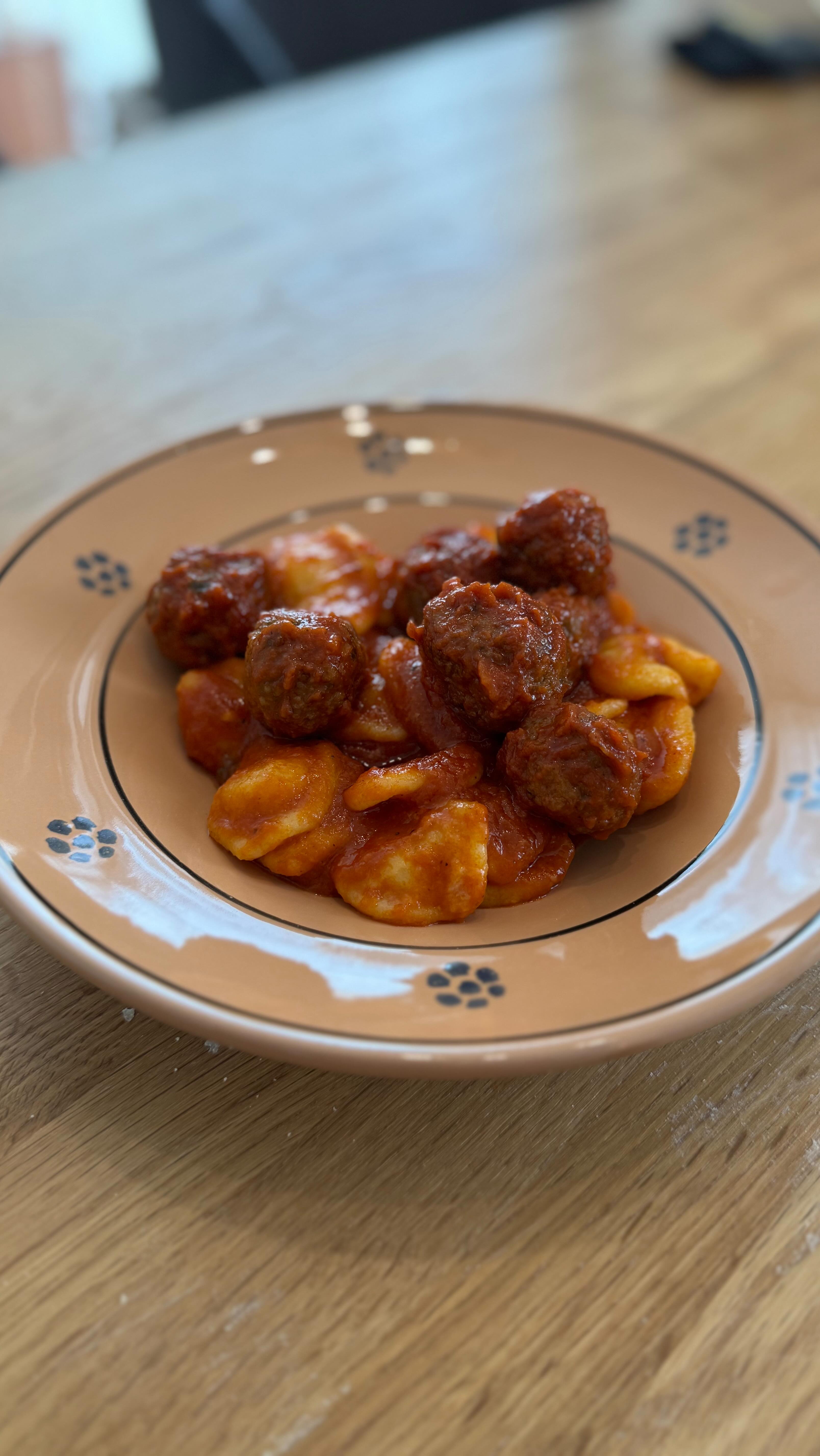 Polpette con orecchiette
Per le Orecchiette:
• 280g di semola
• 130g di acqua calda
Impastare per circa 8 minuti fino ad ottenere una consistenza vellutata. Avvolgere nella pellicola trasparente e lasciare riposare in frigorifero per 30 minuti.
Lavorate il pezzo e realizzate un filoncino dello spessore di circa 1 cm, tagliare dei pezzetti e, con una leggera pressione, tirare verso di sé. Posizionare ogni pezzetto sul pollice e girarlo per formare l’orecchietta.
Per le Polpette:
• 500g di carne macinata mista
• Noce moscata
• Mezzo mazzetto di prezzemolo
• 1 spicchio d’aglio
• Pecorino
• Pangrattato
• 1 uovo
• Sale e pepe
Mescolare tutti gli ingredienti e formare delle palline.
In una padella, scaldare l’olio d’oliva e aggiungere mezza cipolla tritata. Rosolare le polpette su tutti i lati a fuoco medio. Aggiungere 1,2 litri di passata e lasciar cuocere. Chi vuole, può aggiungere un bicchiere di vino rosso. Aggiustare di sale, pepe e zucchero.
Cuocere le orecchiette in acqua salata per circa 5 minuti, fino a quando non sono al dente.