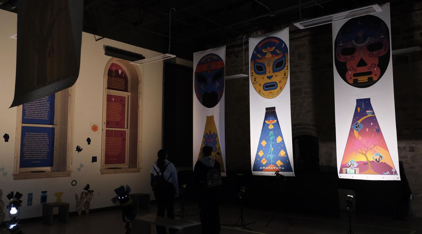 In December, I set up a temporary exhibit of the Lucha Libre Trans Queer Art Espectacular at the Media Arts & Technology Building of New Mexico Highlands University.
I created an installation that showcased the masks, capes, audio stories, and photographs of the athletes. The masks and capes were printed on 44” x 142” canvas paper and hung from the room’s ceiling. I printed the written stories on large fine art paper, which covered the room’s windows. I placed small speakers on each window to play the audio stories, allowing visitors to hear them as they approached. The photographs were displayed on two 85” TV monitors. I designed a mural featuring artwork printed on fine art paper and a wooden cactus I made using the X-tool. The mural unified the artwork, stories, and photographs in the space.