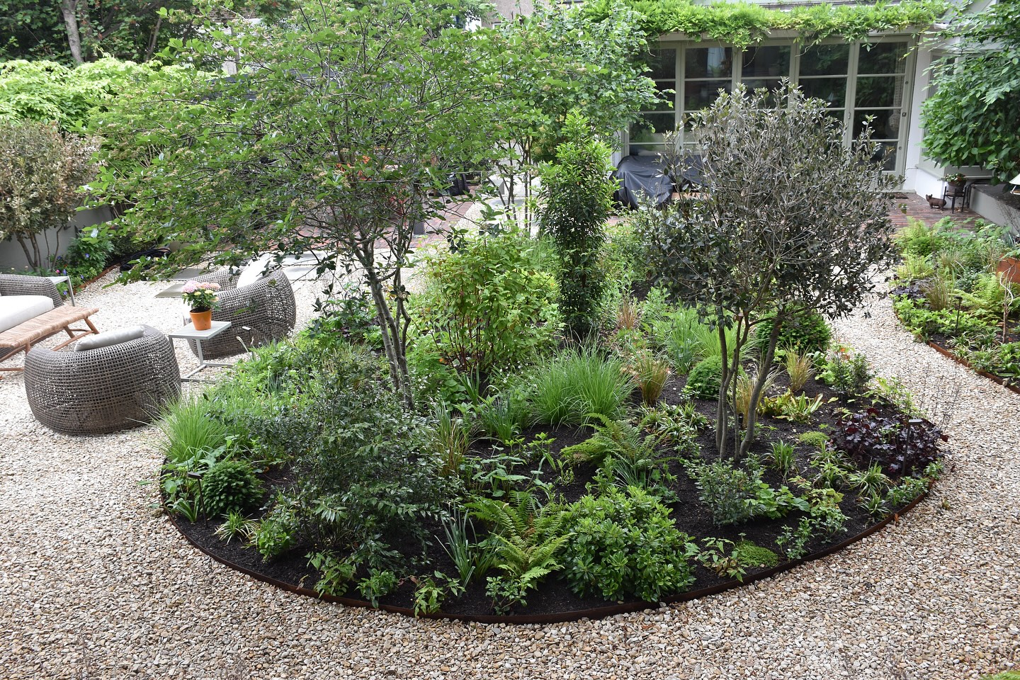 A view over the central planting border in one of my recent garden designs. Multi-stemmed trees – including hawthorn, hazel and ilex – provide structure and seasonal interest, underplanted with a mix of shade-tolerant perennials and grasses.
A curved pathway of Cotswold pebbles from @allgreengb wraps around the border, linking the raised deck (where this photo was taken) to a lounge area for fireside evenings and a separate space for outdoor dining.