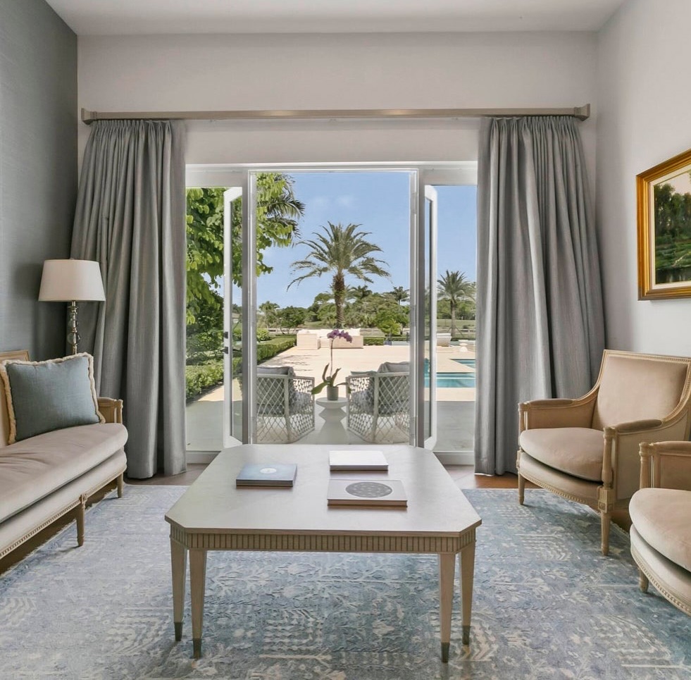 Good morning Sunday! How are you spending your day? 🌴 This master bedroom sitting room exudes elegance and tranquility with a refined, coastal-inspired palette. The space is framed by large French doors that open out to a lush poolside view, letting in abundant natural light and creating a seamless indoor-outdoor connection.
The furnishings are classic and sophisticated, with a neutral base of soft beiges and pale grays. A pair of tailored armchairs and a traditional bench-style sofa are arranged around a pale wood coffee table, all atop a muted blue and gray area rug with a subtle pattern. The pleated drapes in a soft silvery-gray fabric add a layer of luxury and softness to the room.
Decor is kept minimal to highlight the serene atmosphere: a floor lamp with a simple white shade, a few curated coffee table books, and a single piece of framed art on the wall. The view of swaying palm trees and a pristine poolscape beyond enhances the resort-like feel of this relaxing retreat. It’s a timeless and polished space, ideal for unwinding in peace.