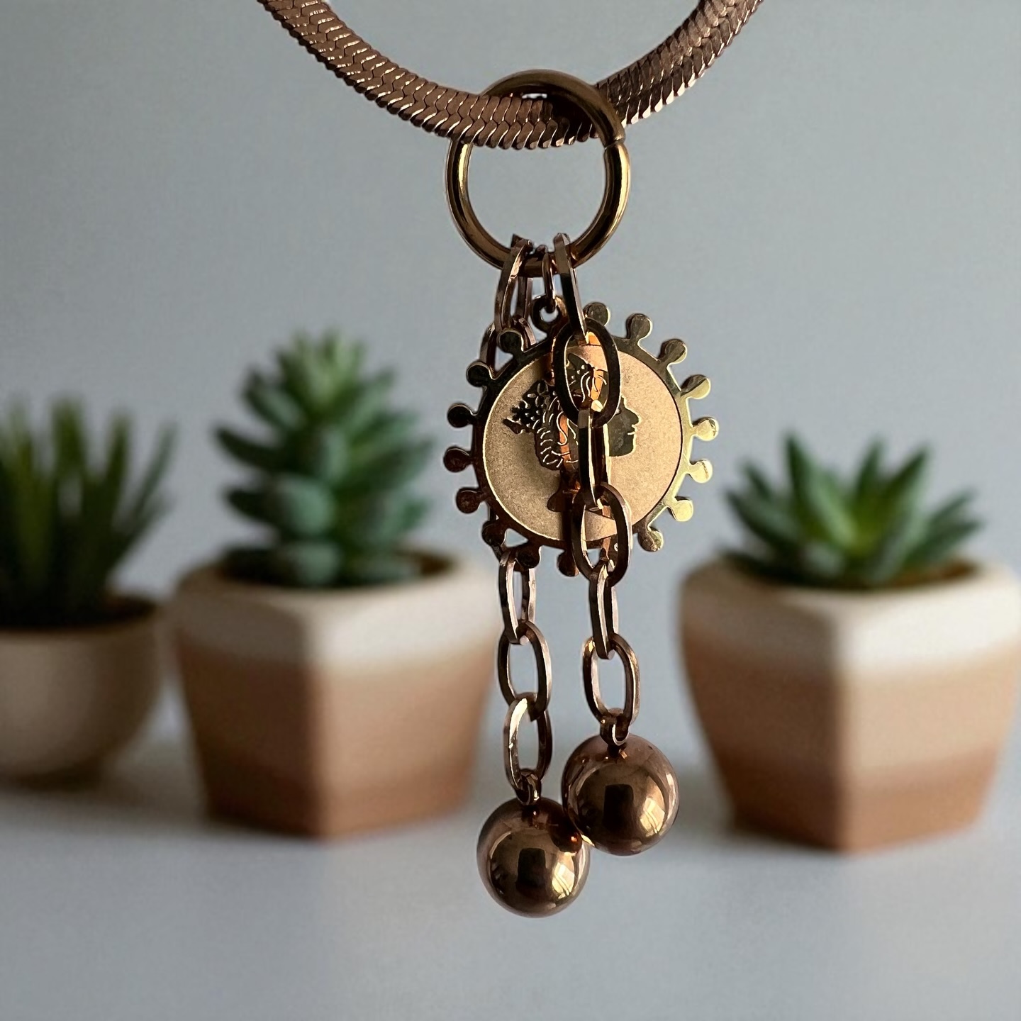 The Roman Coin & Orb Medallion Necklace- Discover a necklace that tells a story of ancient elegance and modern charm. This unique piece features a sleek, rose-gold or bronze-like finish. The pendant is a stunning cluster of three charms: a central, round medallion with an intricate, coin-like relief and a detailed profile of a face. Hanging below are two identical, polished ball charms that add a beautiful, graceful movement. This necklace is a perfect blend of vintage inspiration and contemporary style, making it a versatile statement piece for any occasion, from a casual day out to a more formal gathering.
