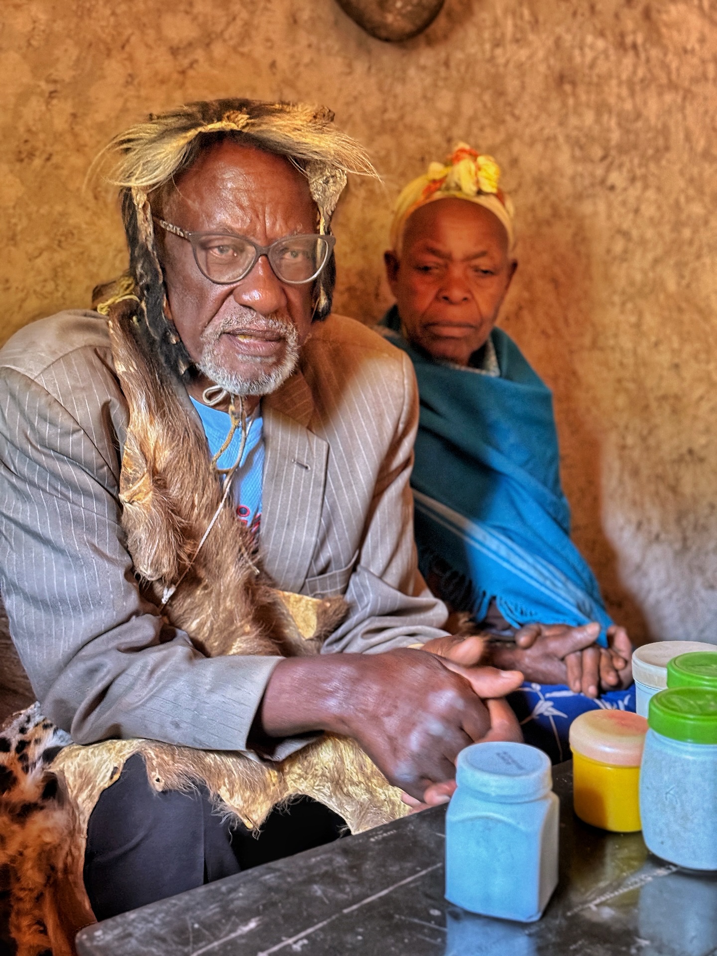 After spending time with our @globallivingston community partners at local schools, health clinics, and farms, I opted for a bit of a different afternoon detour…
I was curious to visit Dr. Sam, a man widely known throughout the Lake Bunyonyi region as a traditional healer and herbalist — and, more pejoratively, a witch doctor.
He’s even been written about in the NY Times, and I was fascinated to learn about his craft, its continued pull, and the relationship between traditional and modern medicine today.
As our boat pulled ashore through tall reeds, we were swarmed by ten of Dr. Sam’s 56 grandchildren, who insisted on holding our hands as we climbed the steep path to his hillside home.
There, we met Dr. Sam, dressed in his usual visitor garb: a jersey, grey blazer, a serval cat pelt, and a furry monkey-hide headdress. He welcomed us into his small mud hut, lined with antlers, horns, and jars of herbs, and began sharing how he treats everything from toothaches and snake bites to mental illness and matters of the heart (yes, he has a love potion, which he said is a best-seller!!).
He demonstrated his two “tests” to see whether he can help a patient—balancing a small horn on a stick, and boiling a bottle of green liquid to see if it changes color. If both fail, he knows they are doomed and suggests they go to a hospital.
Through our translator, we learned that Dr. Sam’s treatments mix spiritual tradition with practical herbalism. Some of his remedies—like teas for arthritis or powders for allergies—are trusted by the community. He accepts payment in chickens, vegetables, or homebrew. And while he can’t cure conditions like HIV, he says his concoctions can “make the virus drunk and weak.”
We met two of his three wives and learned how families still seek his care first, especially for emotional or spiritual ailments or curses.
Our GLI public health colleagues explained that Uganda’s health ministry now partners more with (and tries to impose regulations on) healers like Dr. Sam, recognizing that they are often the first stop for care in rural areas.
A fascinating visit—equal parts surreal, joyful, and thought-provoking. One we won’t forget anytime soon…