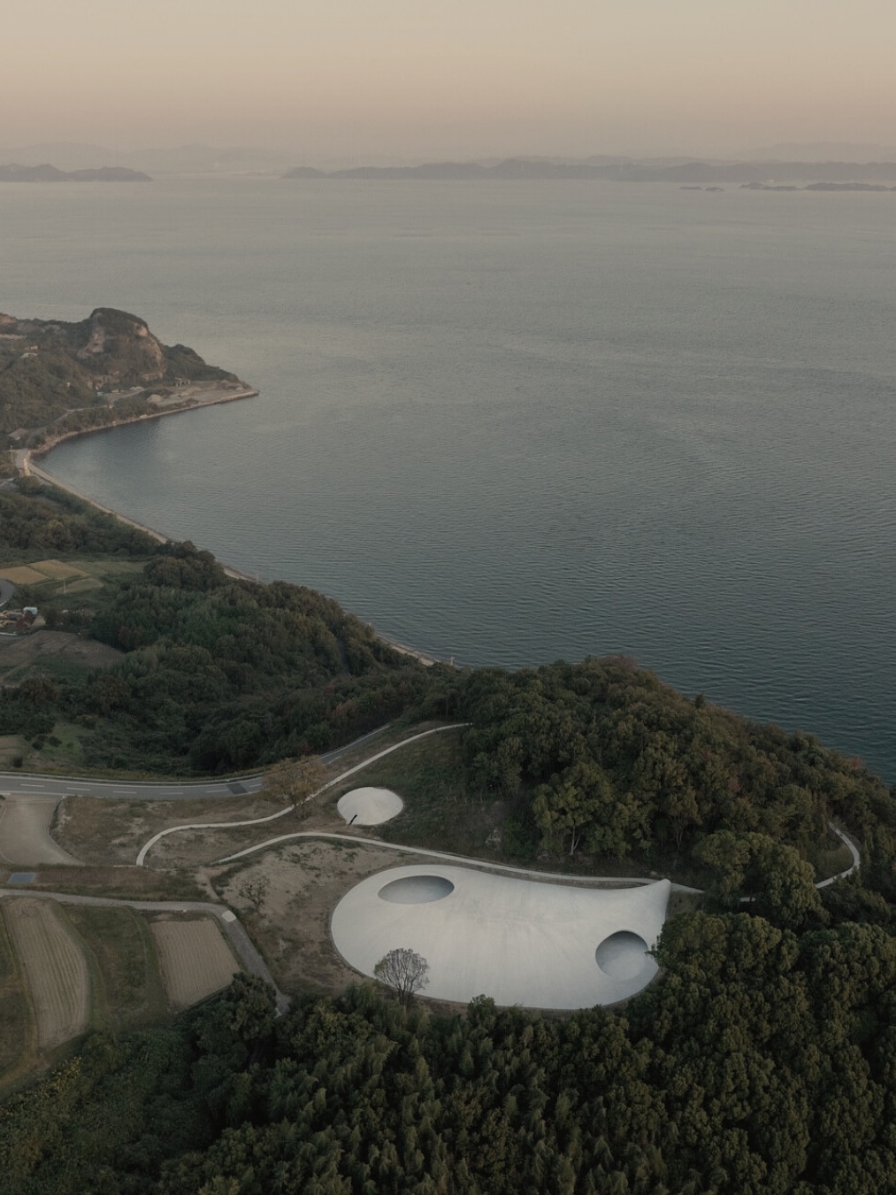 Le plus fascinant dans le Musée d’Art de l’île de Teshima — c’est qu’il n’a ni murs habituels, ni expositions traditionnelles : l’espace est l’œuvre d’art.
Sa coque en forme de goutte d’eau géante se fond doucement dans la terre, comme si elle avait toujours appartenu à l’île. Deux ouvertures circulaires dans le toit — unique « frontière » entre le ciel et l’intérieur — laissent entrer le vent, la pluie, les rayons du soleil et les étoiles. Le musée vit au rythme de la nature : l’eau se rassemble en délicats miroirs, les feuilles glissent lentement sur le sol lisse, et la lumière dessine des motifs mouvants d’ombres.
Conçu par l’architecte Ryue Nishizawa et l’artiste Rei Naito, il est un rare exemple d’architecture qui ne sépare pas l’homme de la nature, mais l’inscrit dans son souffle. Ici, pas de lumière artificielle, pas de climatisation, pas de barrières — seulement le silence, le vent, l’odeur de la mer et le bruissement des feuilles. Chaque instant devient une installation unique, créée par le ciel, la terre et le temps.
#TeshimaArtMuseum #ArchitectureJaponaise #RyueNishizawa #ReiNaito #ArtEtNature #ArtContemporain #Musée #IleDeTeshima #Setouchi #JaponInsolite #VoyageAuJapon #DesignMinimaliste #PoésieArchitecturale #ExpérienceSensorielle #ArtEtArchitecture #VoyageDeLuxe #DestinationUnique
