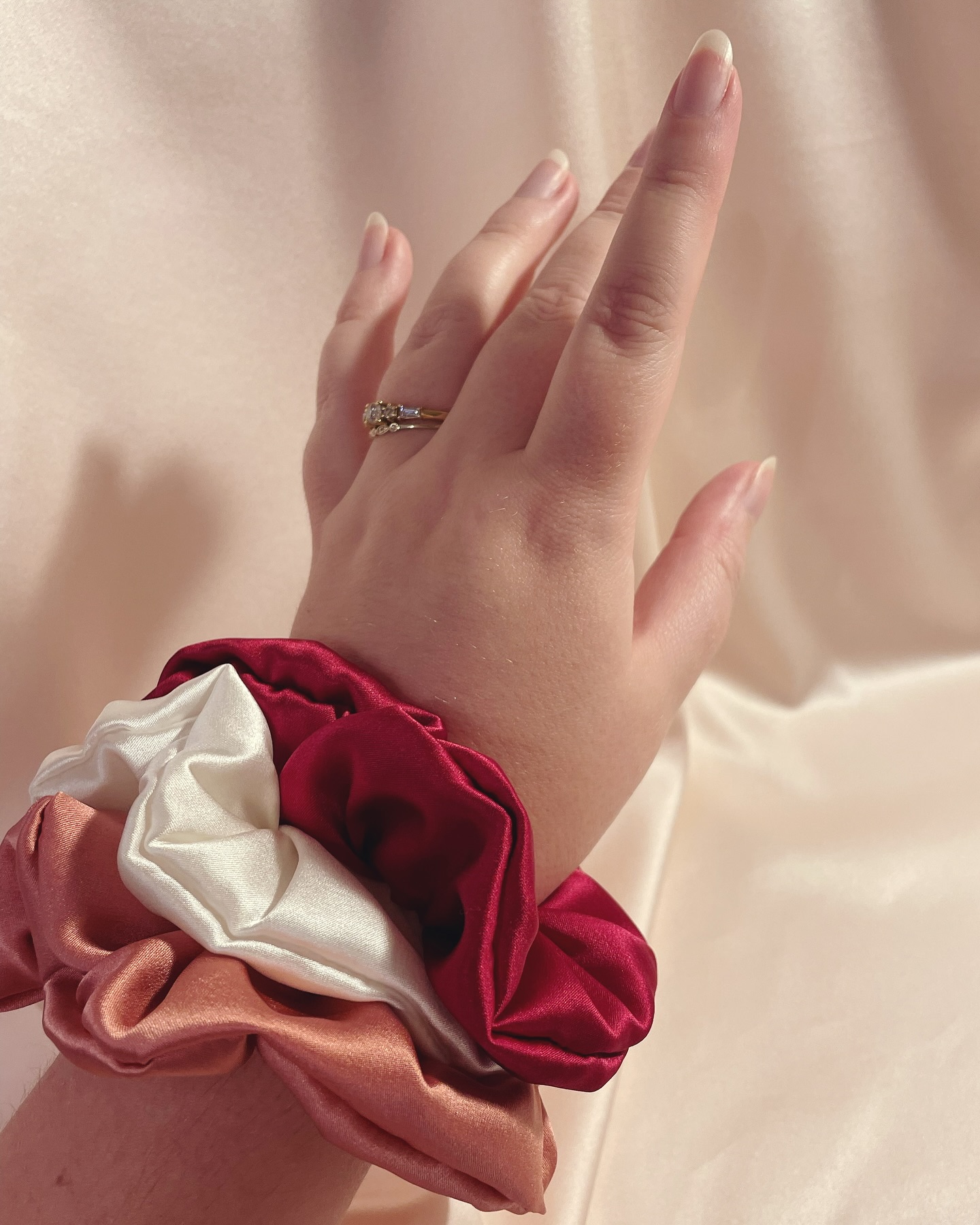 Scrunchie wrist stack- does your scrunchie spend more time in your hair or on your wrist?
Image description- Joās hand with three silk scrunchies in deep red, white and mid pink on her wrist against a pale pink silk background