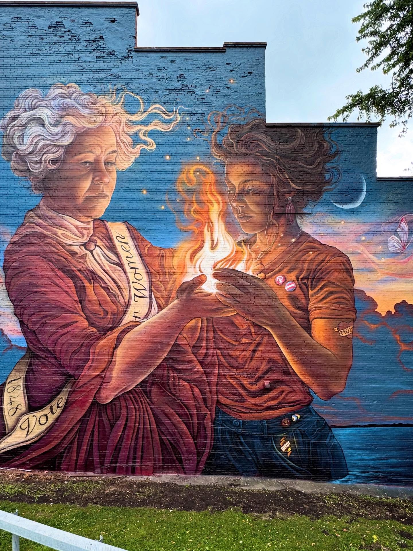 “The Flame Keepers”
A tribute to the enduring fight for women’s rights across generations.
This mural captures an intimate moment of exchange. A suffragette passing a living flame into the hands of a modern woman. The fire represents knowledge, resilience, and the ongoing struggle for justice and equity.
The suffragette’s steady presence honors the women of Seneca Falls who fought to secure the right to vote, while the younger woman receives the flame with reverence and determination, carrying that light forward into a more inclusive future.
Both figures rise from the water, a symbol of rebirth and the roots of Seneca Falls, where the first Women’s Rights Convention reshaped history.
Her sash belongs to the past. Her buttons belong to the present.
And the moths gather in remembrance, for everyone, who gave their life to the cause.
Let’s keep the light burning for all.