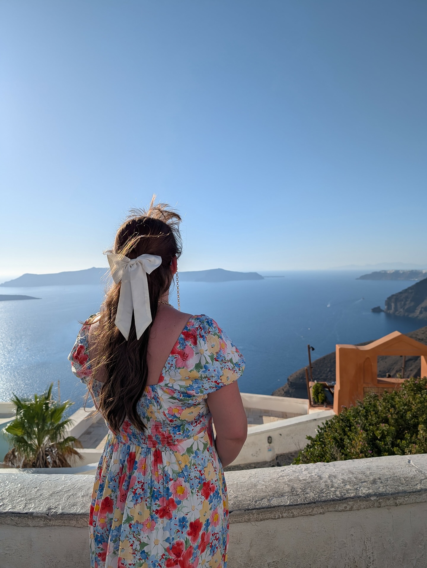 I know it is very much autumn now but please let me have this last summer-y photo!
It was quite windy in Fira so also please ignore whatever my fringe is doing and just focus on the bow 😅 It’s hard to get photos of myself wearing my hair accessories as they are normally at the back of my head and when we go out I often forget.
Image description- Jo, a white woman in her mid thirties is facing away from the camera toward the sea. She is wearing a multicoloured floral dress and a white silk hair bow in her long dark brown hair