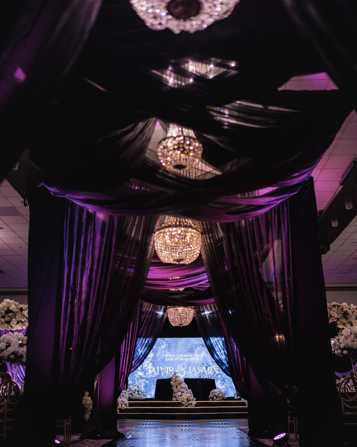 An opulent event entrance illuminated by grand chandeliers, leading to a glamorous ballroom adorned with golden accents and elegant floral arrangements
Design Decor & florals @universaldecor
Venue @ariaconventioncentre
Vinyl / screen Design @designerdancefloorsbc @digitallytailored
Video @liomas14
Our stunning couple @jasminsekhon23 & Jaivir
#WeddingReception #WeddingDecor #ReceptionDecor #LuxuryWeddingDecor #WeddingInspiration #WeddingDesign #WeddingStyling #WeddingDetails #WeddingVibes #eventdécor