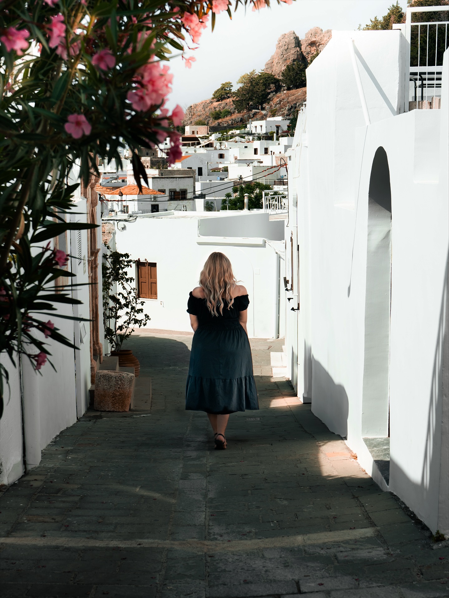 Lindos wirkt auf den ersten Blick fast wie ein Modell aus weißem Karton. Häuser, die sich dicht an den Hang schmiegen, flache Dächer, klare Linien, kaum Schnörkel. Diese Stadt wurde gebaut, um mit Licht zu arbeiten. Mittags reflektieren die Fassaden so stark, dass die Gassen selbst wie Lichtschächte wirken. Sobald man ein paar Schritte weitergeht, ändern sich Schatten und Temperatur, als ob die Architektur den Tag aktiv lenkt.
Auffällig sind die Türen. Viele aus massivem Holz, mit eingelassenen Paneelen und dunklen Griffen, die über Jahrzehnte abgegriffen wurden. Andere in kräftigen Blautönen, die sich gegen das Weiß der Wände absetzen. Man versteht sofort, dass hier nichts zufällig ist. Selbst in den kleinsten Details steckt Handarbeit. Und dann sind da die Höfe, die man nur erkennt, wenn ein Tor offen steht. Steinfliesen in geometrischen Mustern, Tonkrüge, Rankpflanzen, die wie natürlich gewachsene Vorhänge funktionieren und Räume gliedern, ohne sie zu verschließen.
Die Straßen folgen keiner Logik, die man von oben versteht. Sie winden sich, verschmälern sich, öffnen sich wieder zu kleinen Plätzen. Alles wirkt kompakt, aber nie beengt. Der Blick fällt oft auf eine Wand, nur um einen Moment später um die Ecke auf eine offene Treppe zu treffen, die ins Nichts führt und dann doch zu einem Innenraum oder einer Dachterrasse gehört. Es ist eine Architektur, die mit Überraschung arbeitet. Und mit Intimität.
Dass hier Pflanzen so selbstverständlich wachsen, trägt viel dazu bei. Oleander, Bougainvillea und Weinranken bilden ein Gegengewicht zu dem strengen Weiß der Fassaden. Sie weichen es auf, lassen harte Kanten weicher erscheinen und bringen Bewegung in eine Umgebung, die sonst fast abstrakt wäre.
#lindos #rhodos #greece #architecturelovers #mediterraneanstyle #whitetown #oldtownvibes #stonepaths #woodendoors #courtyards #islandlife #travelphotography #fokusfernweh