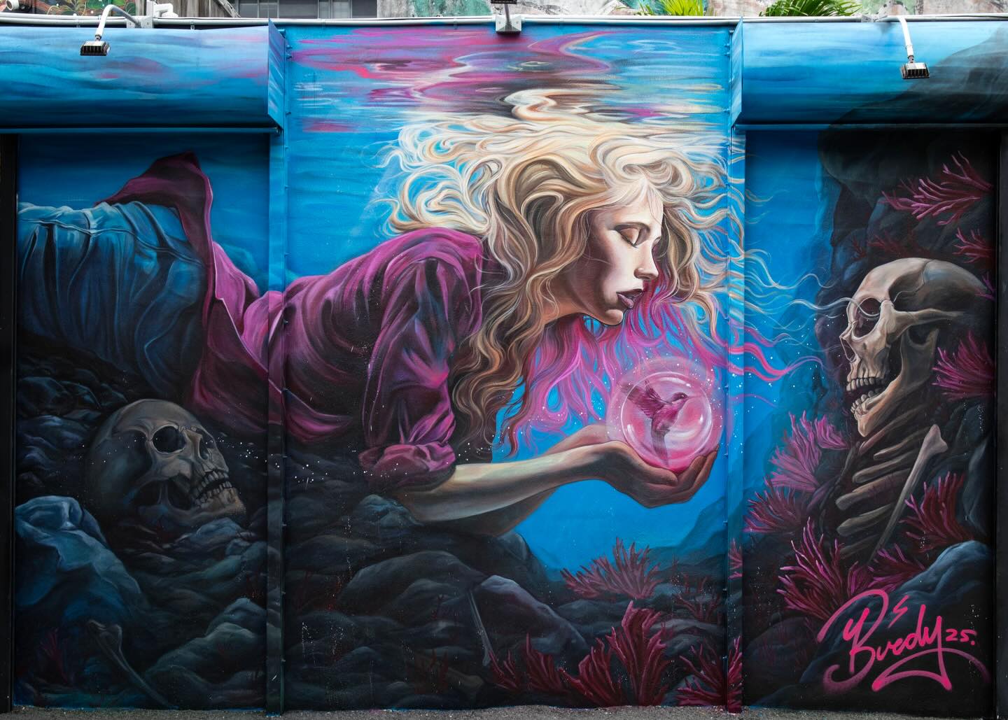 I know you shouldn’t always bare your soul to the world, but when it comes to my work these days, it’s hard not to. This piece is created for the two people who made me, not just physically, but through passion, fortitude, relentless determination, and a deep desire to be good.
In searching for connection with my late parents, I’ve found myself diving deeper into the world I’m building. One where strong women protect, harness power, and deliver truth. It felt bold to create a self-portrait in a place like Wynwood Walls, but no other image could fully express what it means to be “Only Human”.
I present my latest and final painting of the year:
“Bringing the Light Home”
Bringing the Light Home is set underwater, a place that represents the quiet and pressure of loss. The central figure is offering a glowing hummingbird to a skull, a way of trying to reconnect with her mother who recently passed.
Her father’s skull rests nearby, symbolizing an older grief that has become part of her foundation. This act of giving light to the dead is both impossible and deeply human. It is the instinct to keep loving when there is no one left to receive it. The piece reflects what it feels like to live with grief, how it slows you down, makes everything heavier, but still asks you to keep going. Even in that stillness, there are moments of light trying to break through. The mural is not about drowning. It is about learning to breathe differently. It is about the resilience of love that refuses to disappear. Bringing the Light Home is a reminder that what we have lost is never gone. It simply waits for us to find new ways to carry it forward.
Thank you to everyone that made a safe space for this creation @wynwoodwallsofficial
Shout out to @marthacoopergram @nikakramer @photographygoat_ @zuzu_grateful @oliverosisabela for the incredible documentation and laughs. Till next time 💕