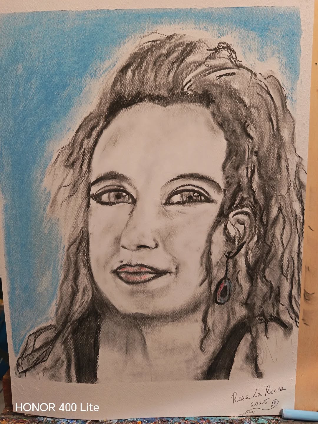 #charkcoalpotrait
🌑 Gestione delle Ombre e del Chiaroscuro nel Ritratto
Il carboncino è un medium ideale per l'espressività e per la rapidità nella creazione di un contrasto drammatico. Nel ritratto di Eleonora, l'artista ha impiegato il carboncino in modo mirato per definire la forma e l'emozione:
1. Definizione della Forma (Contorni e Massa)
Linea Spessa: La linea di contorno (ad esempio, lungo il profilo del naso, l'arcata sopracciliare, la linea della mandibola e, in modo molto evidente, nei capelli) è spesso calcata e scura. Questo non solo separa la figura dallo sfondo ma ne rafforza la presenza e la solidità.
Ombreggiatura Esemplare sui Capelli: I capelli sono l'area con la maggiore densità di carboncino. L'artista ha utilizzato linee spesse e curve per suggerire riccioli e onde, riempiendo le aree più interne con un nero fitto (le ombre proprie), e lasciando il bianco della carta o un grigio leggero per i punti luce, conferendo grande volume e movimento alla capigliatura.
2. Rilievo e Rotondità del Volto
Ombre Medie (Mezzitoni): Per dare rotondità al volto, il carboncino è stato sfumato in tonalità di grigio. Queste ombre non sono nette ma diffuse, e definiscono il volume delle guance, la zona sotto le labbra, i lati del naso e l'area sotto l'arcata sopracciliare.
Questo uso attento dello sfumato (mezzitono) è cruciale per passare gradualmente dalla luce intensa (il bianco della carta) all'ombra più profonda.
Punti di Ombra Profonda: Le ombre più scure (punti di massimo contrasto) sono posizionate strategicamente per definire i recessi:
Sotto l'orecchino.
Nelle palpebre superiori e nelle pieghe dell'occhio, che aggiungono profondità allo sguardo.
Nelle fosse nasali e immediatamente sotto il labbro inferiore, per dare proiezione al naso e alle labbra.
3. Contrasto Drammatico (Luce e Sfondo)
Luce di Provenienza: Sebbene non esplicitamente definita, la luce sembra provenire dall'alto e frontalmente o lateralmente, lasciando le aree come il collo e i lati del viso in una penombra morbida.
Contrasto Sfondo/Figura: L'uso del pastello azzurro come sfondo piatto e brillante serve a isolare e far emergere la figura (soprattutto i capelli scuri) e