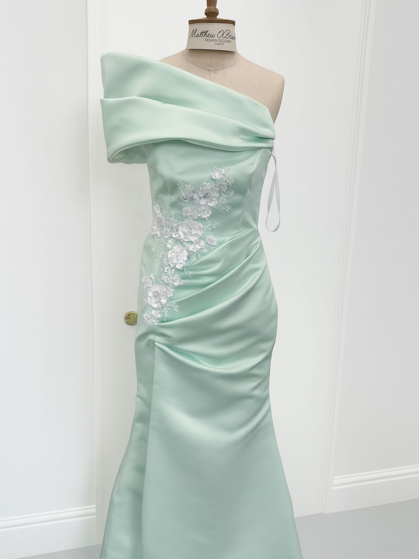 A mint satin couture gown featuring a sculpted off-shoulder neckline, hand-applied white floral appliqué, and soft ruching that flows into a sleek, floor-length silhouette.