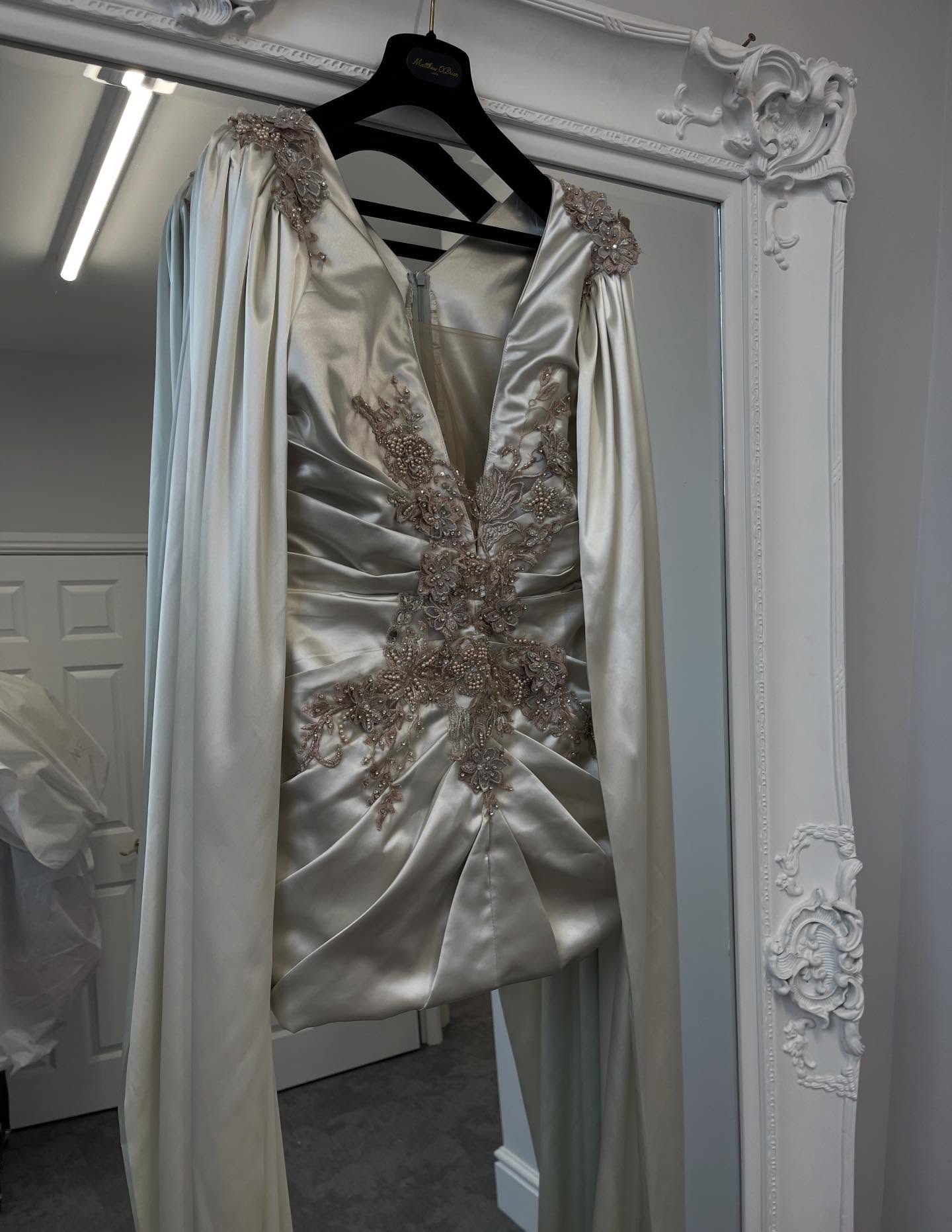 Elegant gown featuring intricate pleats and embellishments with a dramatic long cape sleeves
