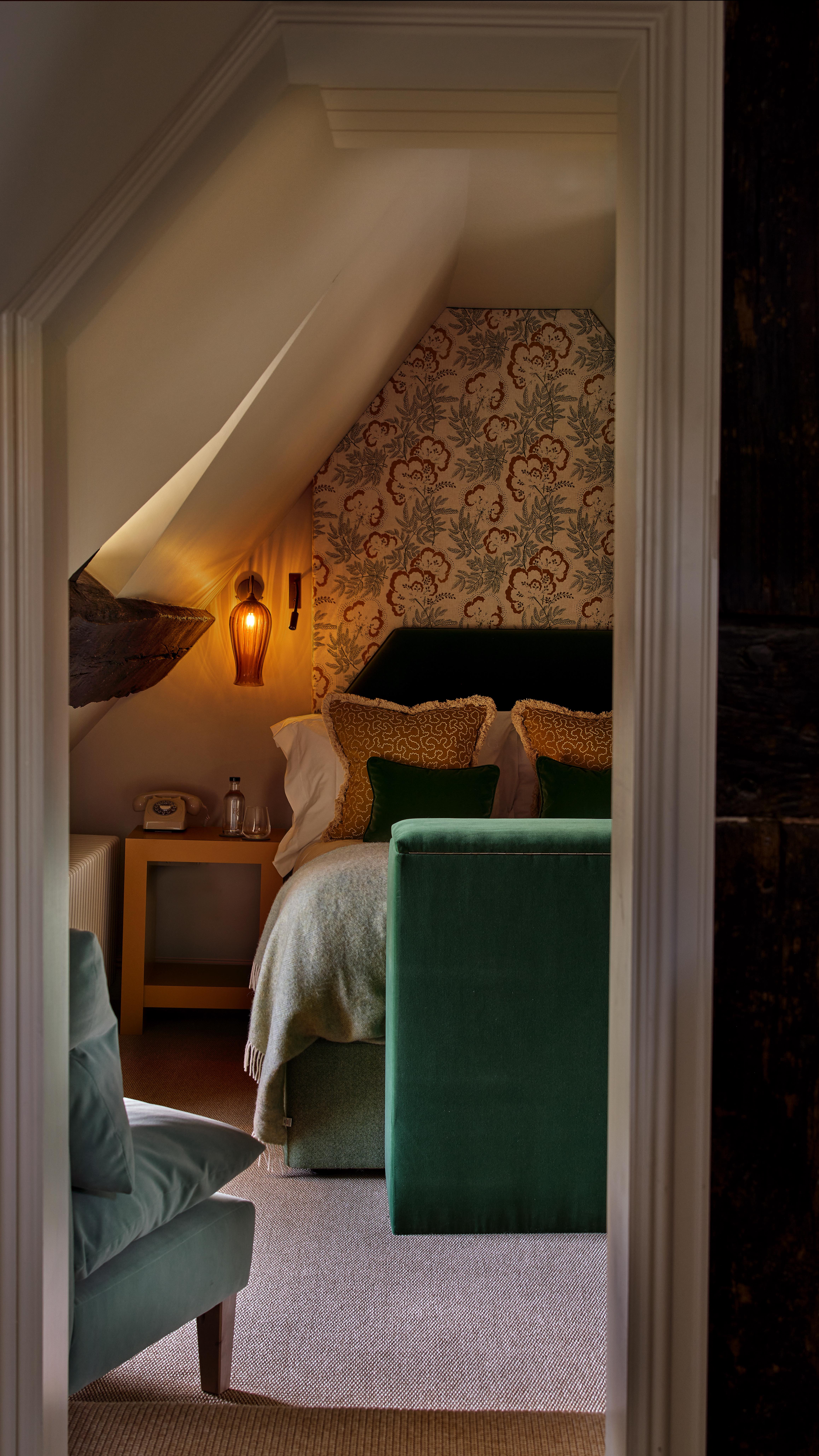 Softly upholstered walls and a cosy double bed make The Eaves perfect for quiet moments.
The en-suite features bespoke tiles and a two-person shower, while the window frames a view of Broadway high street beneath original timber beams.