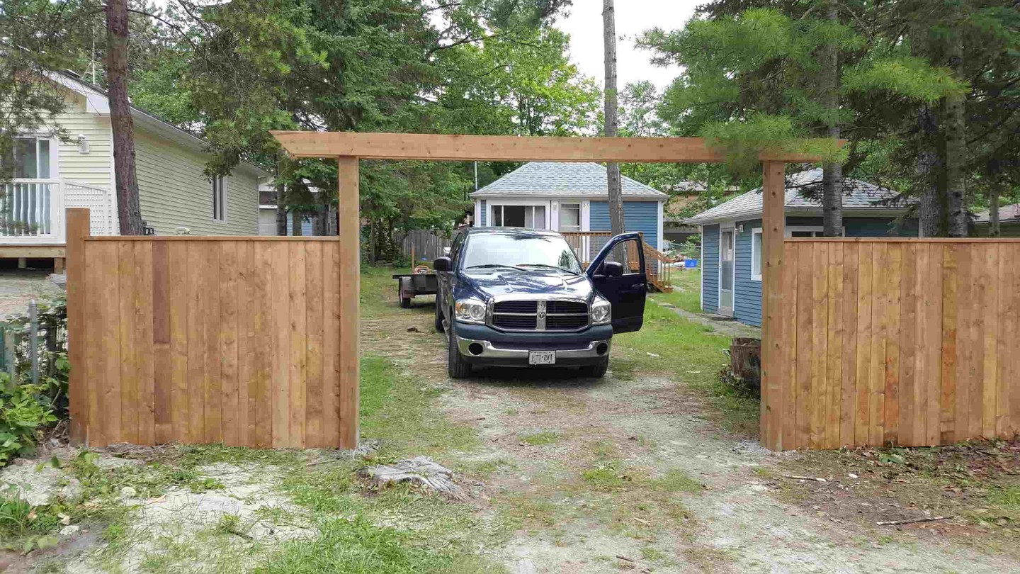 We finished up this custom driveway gate and fence for a property surrounded by tall trees and classic cottages.
The solid wood posts and overhead beam give the entrance real presence, while the vertical planking ties everything together with that rustic, natural look.