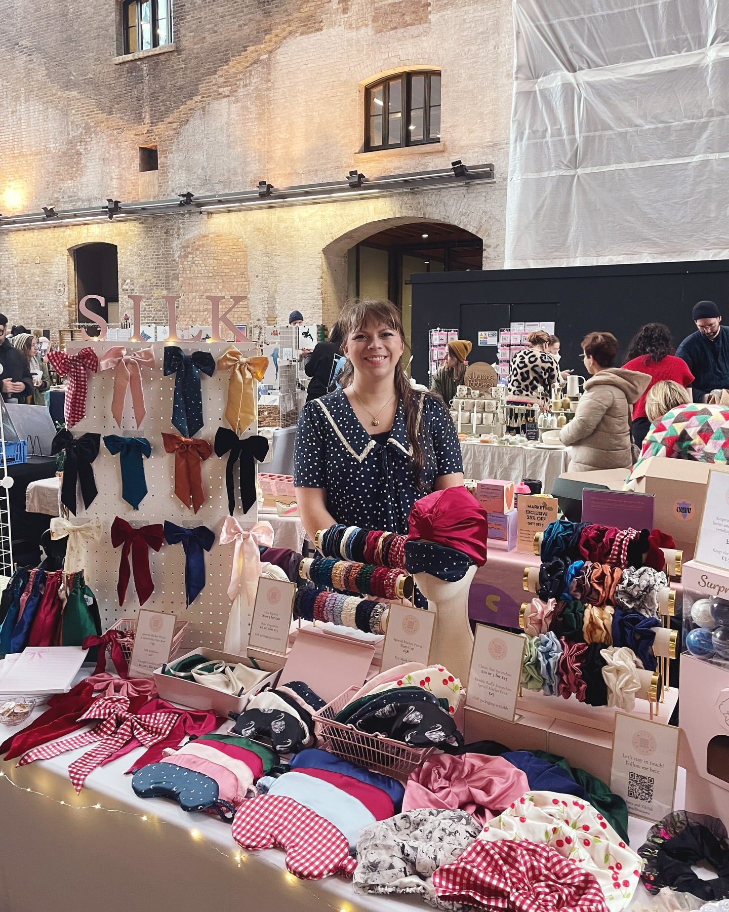 I will be back at Kings Cross with @craftyfoxmarket this Sunday for my last Christmas market of 2025! The scrunchie vending machine will also be with me
I had the loveliest moment this past Sunday when two girls each bought a silk ribbon for their hair and one went to tie it into the other’s hair straight away. I helped teach them the best way to tie a hair ribbon so it hangs evenly and their excited reaction really was the best moment of the day! 🎀
I also got to buy some presents and had my glamourous market assistant with me again 🤭
Image description – 1. Jo, a white woman in her mid thirties, is stood behind a market table filled with her silk accessories including hair bows, eye masks, scrunchies, sleep caps and a scrunchie vending machine
2. A close up of the table
3. Another close up of the table
4. A third close up of the table
5. The same table but with Raff, a half Italian man in his mid thirties, standing behind it