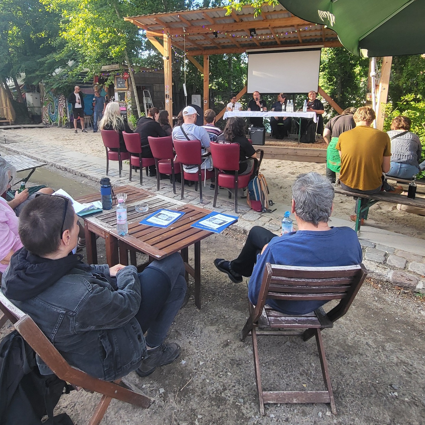 eit dem 19. Jahrhundert hat sich im Zusammenhang mit der Entstehung der modernen Großstädte und dem damit verbundenen sozialen Elend ein neues literarisches Genre herauskristallisiert – die „Urban Poetry“. Sie reflektiert die Erfahrungen der urbanen Existenz und sieht es dabei oftmals als ihre Funktion an, „den Finger auf den wunden Punkt“ zu legen. Im Rahmen einer niedrigschwelligen Veranstaltung in der „Teepeeland“-Siedlung sollte den TeilnehmerInnen am 31.05. ab 19 Uhr das Spektrum der Urban Poetry in seiner ganzen Vielfalt dargelegt werden. Die Vortragenden waren die indische Soziologin/Aktivistin Khushboo Jain, die Germanistin/Kulturmanagerin Bettina Henningsen und die Niko Rollmann.
Das Spektrum der vorgetragenen Gedichte umfasste „Klassiker“ von Tucholsky, Ringelnatz und Kaléko, Werke bekannter Nachkriegsliteraten wie Brinkmann und Linton Kwesi Johnson, gegenwärtige indische DichterInnen und auch einen von Künstlicher Intelligenz (KI) erstellten Text. Besonderes Interesse erweckten die Texte aus Indien, die u.a. die jüngsten blutigen Ereignisse im Kontext der Kaschmir-Region thematisierten.