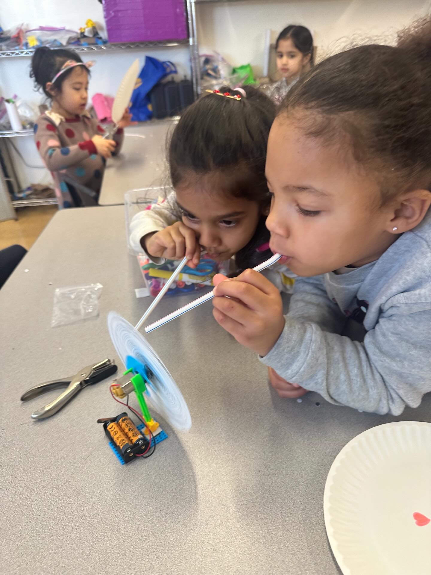 Kindergarteners build a “siren disk” by attaching a paper disk with punched holes to a small fan, then aim a straw at different rings of holes and blow to create tones. By changing which holes they use and how fast the disk spins, they hear how pitch changes and connect it to vibration frequency in sound waves.