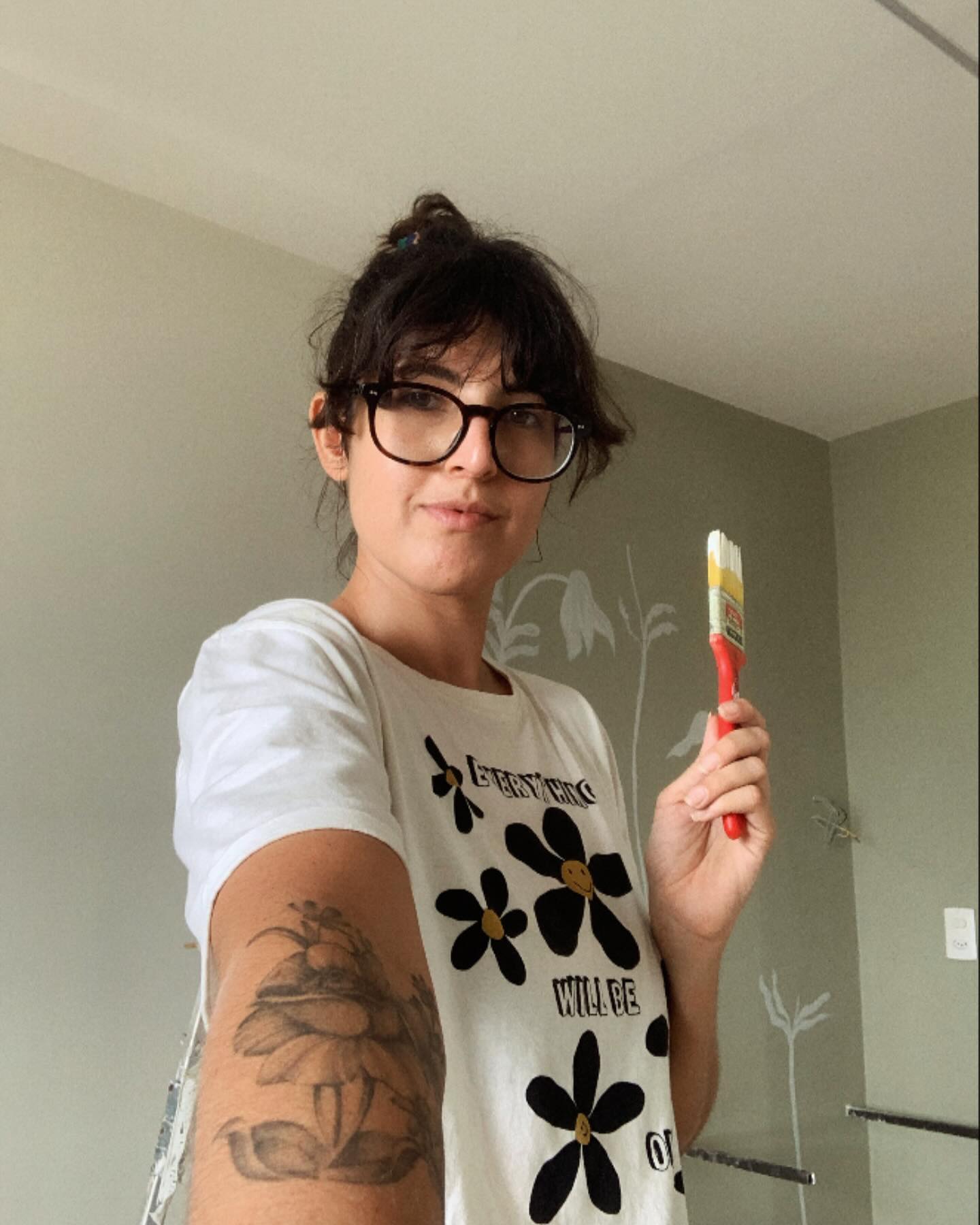 Nesta imagem vemos uma artista, com seu pincel sujo de tinta na mão esquerda, em sua nova versão “sem medo de paredes” e com sua blusa de flores felizes com os dizeres “everything will be ok” 👌🏼🪻🌼