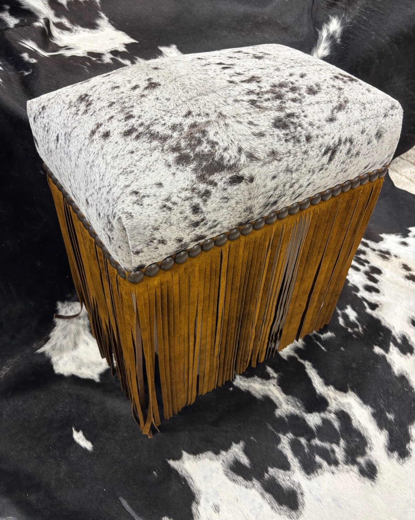 This square ottoman features a spotted gray-and-white cowhide top with bold natural patterns, edged in brass stud trim, and dramatic long tan suede fringe hanging from the sides for a boho-western vibe. Perfect as a footstool or accent seat.
#CowhideOttoman #FringeOttoman #WesternBoho #BohoWestern #CowhideFurniture