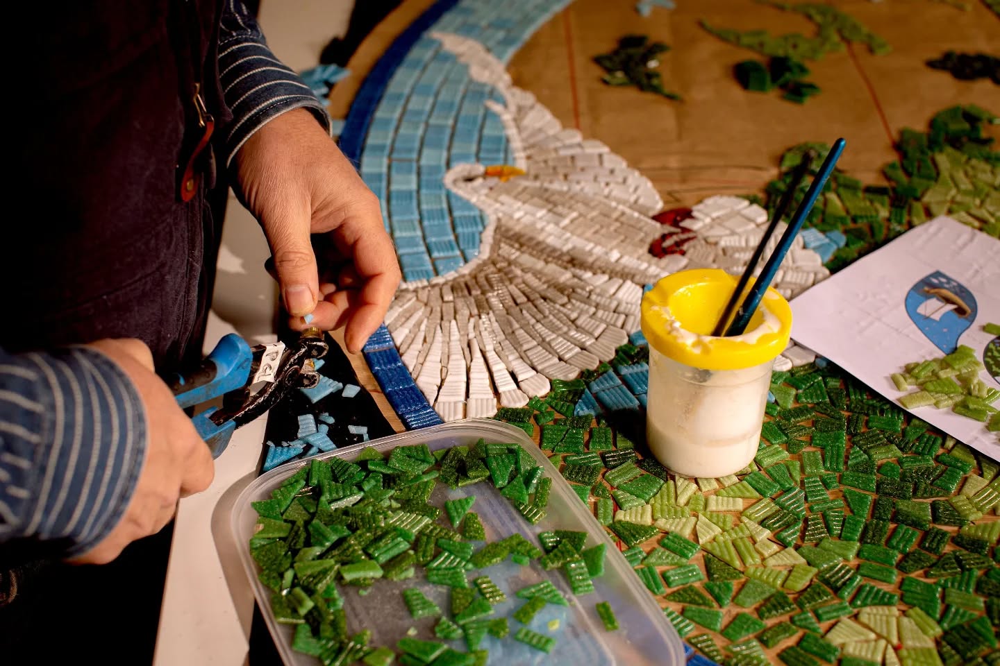 We have been working away on some incredible projects lately, this particular piece is for Hever CofE Primary School in Kent. The design features wildlife and is part of a large mural for the school's hall.
We had the pleasure of having Saul 📸 @hansaulo come and photograph us while we were completing this project in our studio.
This photo shows Paul's hands cutting the blue glass tiles that will be placed around the flying dove. In the foreground is a tray of cut green glass and a glue pot.
#glassmosaic #inthestudio #ihavethisthingwithtiles #themosaicstudio #mosaico