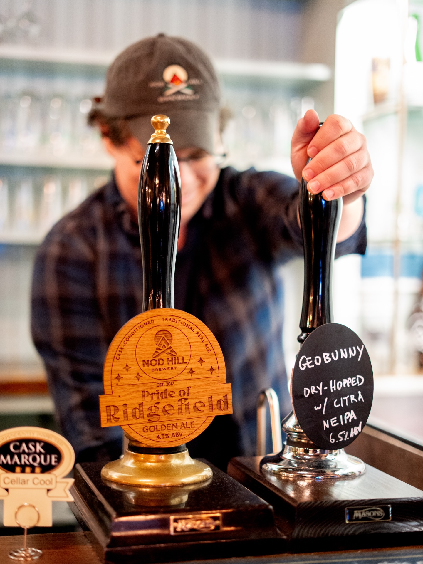 At Nod Hill, we are proud to offer English-style cask ale. We are also proud to be one of less than 20 breweries in the US with Cask Marque Accreditation.
But why are cask ales so special?
- Natural carbonation = less gas, gentler bubbles, easier drinking
- Cellar temperature = more expression of flavor and aroma
- Venting = slight oxidization that subtly changes the beer’s flavor over the days each cask is pouring
The process of creating cask beer is also quite distinct. When beer is finished fermenting but has not yet been carbonated, we transfer it to a cask along with a small quantity of sugar and yeast. This kicks off a secondary fermentation process where the yeast consumes the sugar and creates CO2 as a byproduct.
Once the beer becomes naturally carbonated, we place the casks in a temperature-controlled space (around 50-55°F) and vent the tops of the casks to allow excess CO2 to be released. Then they are connected to a beer engine & hand pump to be served for your enjoyment.
Along with our classic English pub styles, we are excited to offer a cask version of Geobunny at this time. Stay tuned for some fun & interesting iterations of our flagship IPA on cask to complement our more traditional offerings!
#nodhillbrewery #cask #caskale #ctbrewery #ridgefieldct