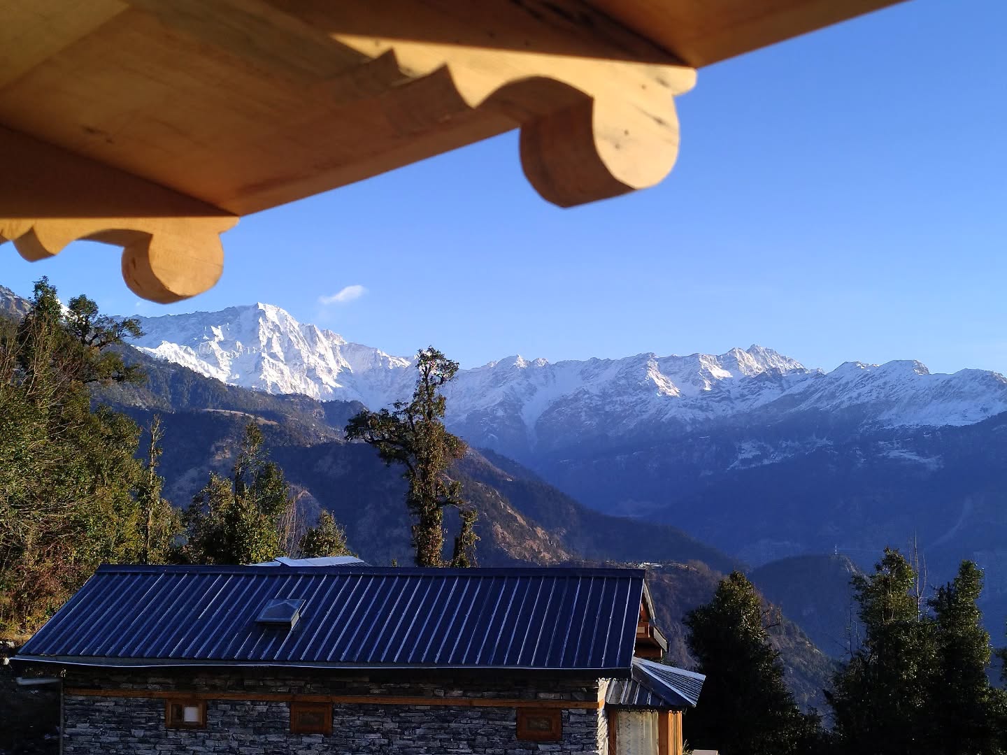 Standing beneath the carved wooden eaves, the snow-covered peaks rise in the distance as if they’re touching the sky. A small stone-roofed house rests quietly below, surrounded by layers of dark green forests and rolling mountain ridges. There’s a deep stillness here—no city noise, no rush—just crisp air and the soft glow of morning light turning the white summits golden. It’s more than a view; it’s a pause, a reminder that nature moves at its own steady rhythm. In this silence, time slows down, thoughts clear, and the heart feels a little lighter..
#himvanliti #natureretreat #naturescape #kumaon #uttarakhand