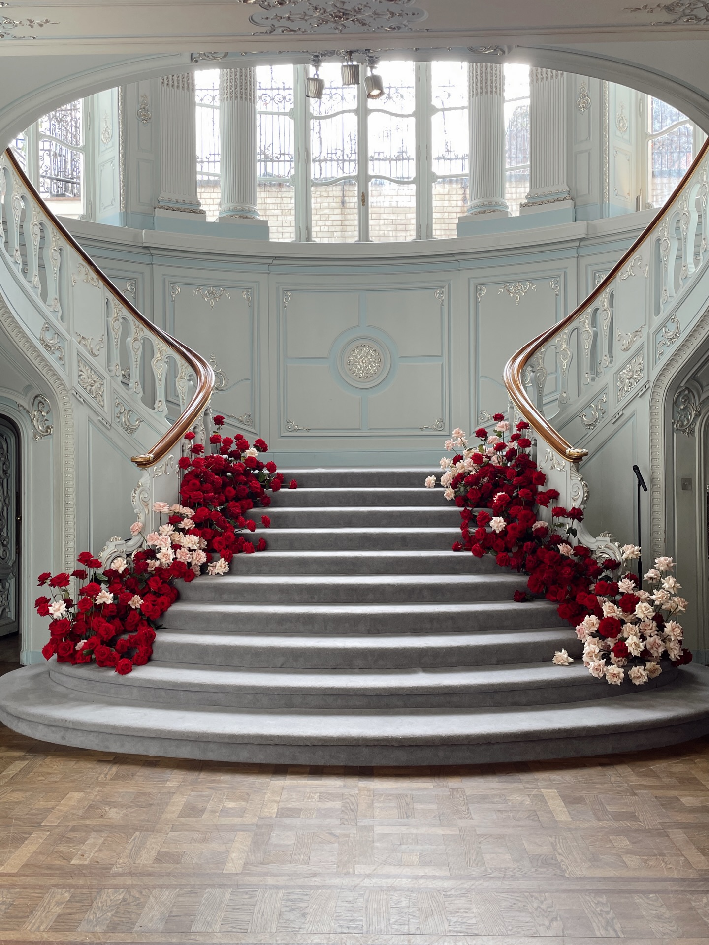 The brief: create a moment of arrival.
This sweeping staircase already carries so much presence. The soft blue panelling, the curve of the bannisters, the height and light pouring in from above. Rather than competing with the architecture, I wanted the flowers to feel as though they were growing into it.
We designed low, abundant clusters of red and blush roses that spill organically from either side, framing the steps without overwhelming their shape where the bride and groom would stand and say their vows. The deeper reds add richness and drama, while the softer pinks lift and soften the palette, a balance of bold and romantic 🌹