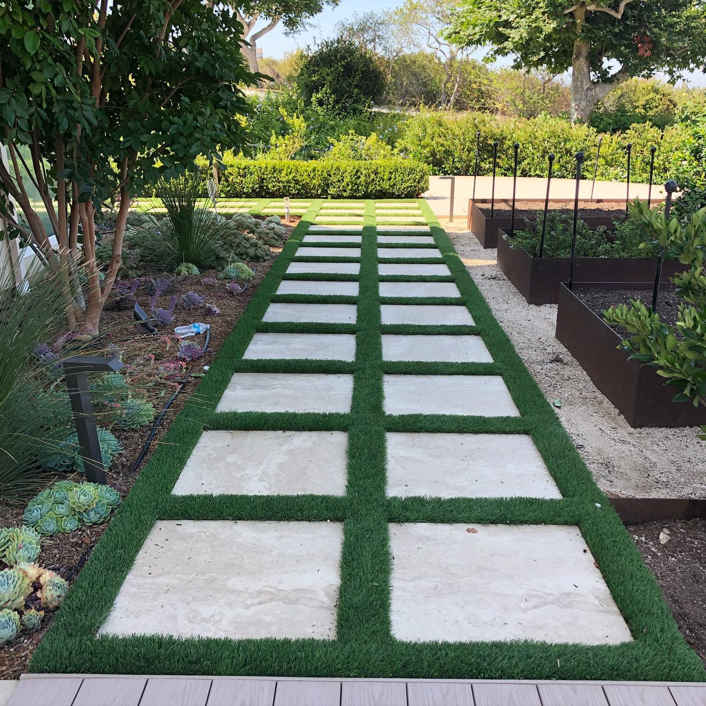 Up ahead on your right, raised garden beds and wood deck. We love where this lush green turf and square paver combo leads✨🌱