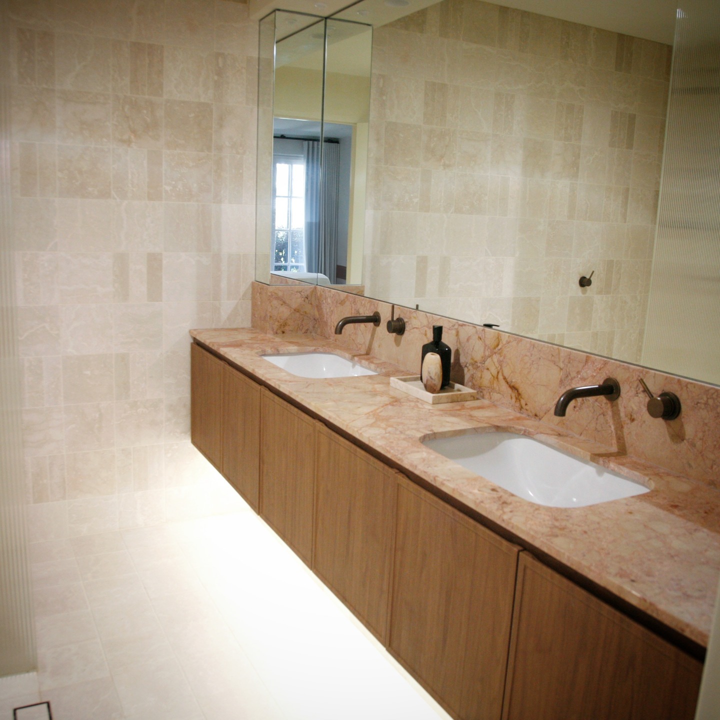 For our Kirribilli Terrace project, the bathroom was conceived as a calm, immersive retreat where materiality leads the conversation. Veined blush marble wraps the vanity like a sculptural plane, its organic patterning bringing warmth and depth against a backdrop of softly tonal marble tiles.
The composition is intentionally restrained. A floating timber joinery unit grounds the room with natural texture, while bronze wall-mounted tapware punctuates the palette with a subtle, graphic contrast.
A mirrored column introduces verticality and reflection, catching light from the adjoining space and extending the room beyond its boundaries. The result is a bathroom that feels both refined and deeply tactile, an intimate space designed to be experienced.
#SydneyInteriorDesigner #LuxuryInteriorDesign #InteriorDesignerSydney #HighEndInteriors #BespokeInteriors #LuxuryBathroomDesign #BathroomDesignInspiration #ArchitecturalInteriors #InteriorArchitecture #DesignerBathrooms #CustomJoinery #StoneInteriors #TimberJoinery #KirribilliInteriors #SydneyHomes #LuxuryHomeDesign #ResidentialDesign #CuratedInteriors #ElevatedInteriors #InteriorDesignStudio #TheEscapistandCo #HomeTransformation