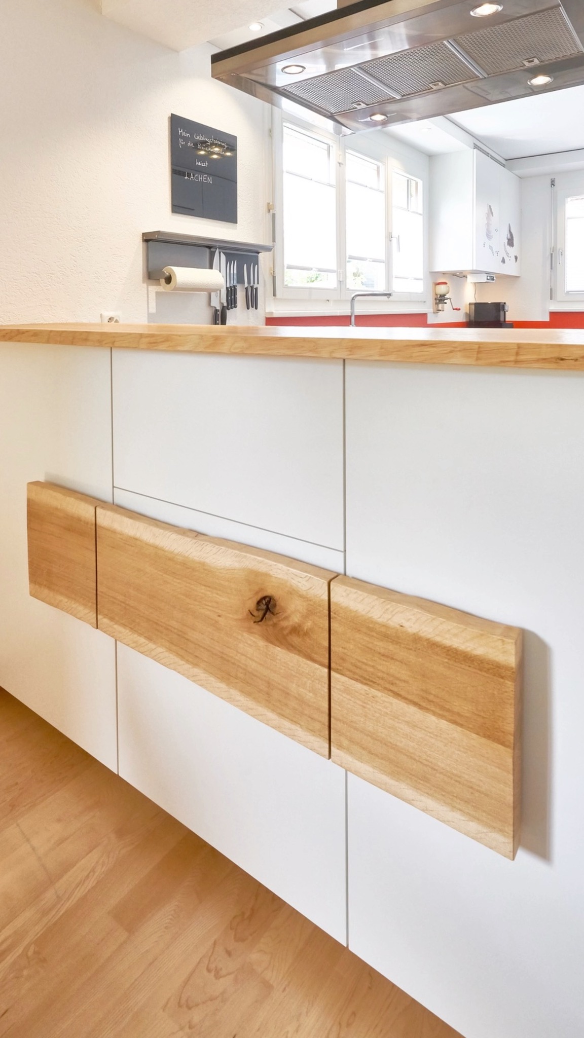Diese Küche zeigt, wie ruhig und klar eine Gestaltung wirken kann.
Weisse Fronten, eine durchgehende Arbeitsfläche und die rote Rückwand setzen bewusste Akzente. Ein besonderes Detail ist das Sideboard zum Essbereich. Die Holzfront greift das Material der Abdeckung auf und verbindet Küche und Wohnraum auf eigenständige Art.