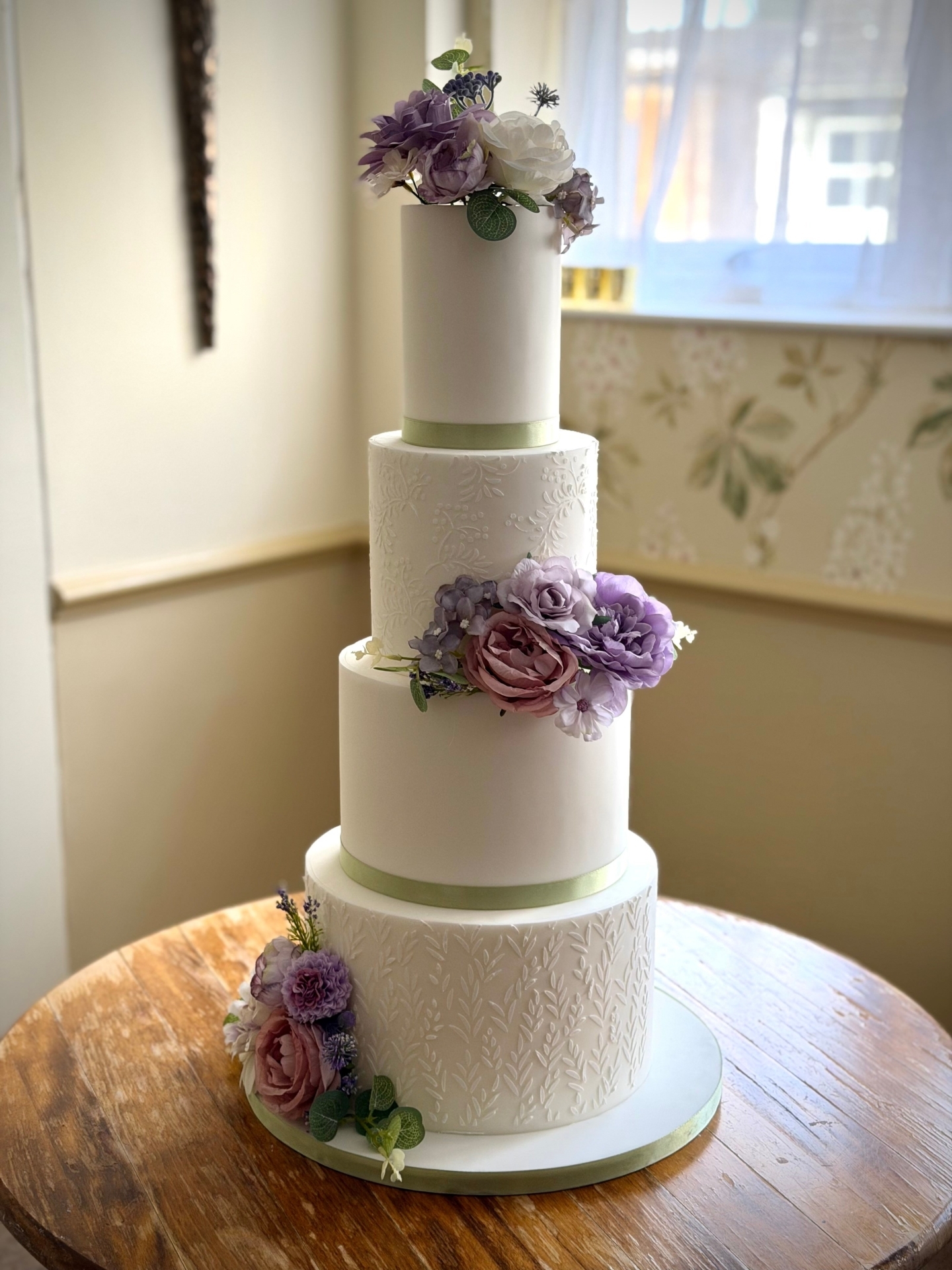 Soft, romantic details with a timeless feel.
This four-tier wedding cake was created as a showcase design, featuring delicate embossed textures, soft sage ribbon accents and beautiful faux florals in lilac, blush and soft ivory tones.
Faux flowers are a lovely option for couples who want the look of florals on their cake while keeping the design lightweight, neat and perfectly positioned throughout the day. They also allow the colours and arrangement to be planned exactly to complement the overall wedding styling.
Elegant, understated and designed to feel effortless.
Enquiries for 2026 and 2027 weddings are now open via the website.
www.kimberlyjaynecakedesign.co.uk
📍Swindon
Serving Wiltshire, the Cotswolds and the South West
#luxuryweddingcake
#weddingcakeuk
#cotswoldswedding
#wiltshirewedding
#bespokeweddingcake