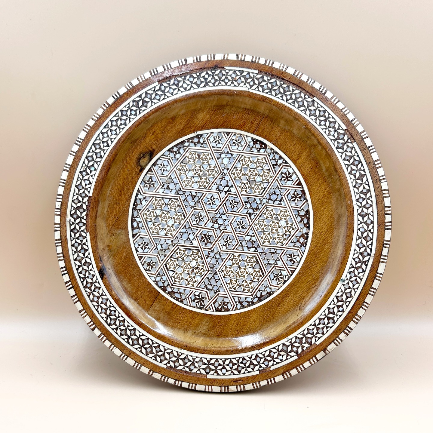 🟢 DOSTĘPNY | Zjawiskowy talerz, Syria, Damaszek, lata 80. XX w.
Okrągły talerz z drewna orzechowego, zdobiony bogatą intarsją z masy perłowej, drewna i emalii.
Centralną część wypełnia precyzyjna kompozycja geometryczna oparta na gwiazdach i wielokątach, charakterystyczna dla sztuki islamskiej i tradycji Damascus work. Szeroki rant otacza dekoracyjna bordiura z powtarzalnym wzorem geometrycznym, podkreślająca symetrię całości.
Spód pozostawiony gładki, eksponujący naturalne usłojenie drewna, co jest typowe dla tego typu wyrobów.
Drobne odpryski na obrzeżu talerza zw. z wiekiem. Oferujemy stelarz do wieszania na ścianie.
Śr. 26 cm
195 PLN + 15 PLN